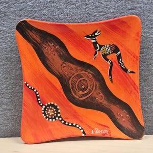May include: Square ceramic dish with an Aboriginal art design. The dish features a kangaroo, a brown and black swirl, and a dotted line on an orange background. The artist's signature, "L. Rosete", is visible.