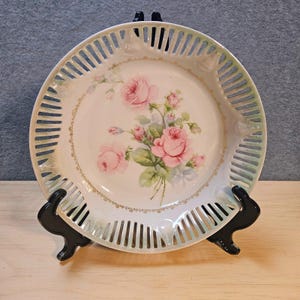 May include: A round porcelain plate featuring a central floral motif of pink roses with green leaves. The plate has a decorative openwork lattice border with a subtle green tint and a thin gold-colored trim encircling the central rose design.