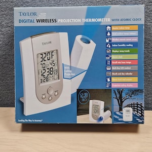 May include: A white and silver Taylor digital wireless projection thermometer with atomic clock. The display shows the temperature in Fahrenheit, humidity, time, and date. Features include indoor/outdoor temperature, humidity reading, and a projection ceiling display.