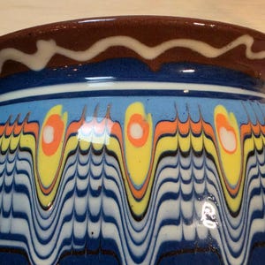 May include: A ceramic bowl featuring a dark brown rim with a white wavy line. The body displays a vibrant marbled pattern with vertical blue, yellow, and orange teardrop shapes layered over a series of fine, horizontal blue and white wavy lines.