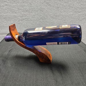 May include: A curved wooden wine bottle holder with intricate pyrography patterns holding a dark blue glass wine bottle. The bottle features a white label with black text and a barcode. The wooden stand has a smooth, polished finish.