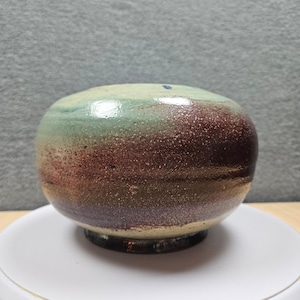 May include: A handcrafted ceramic bowl with a rounded shape. The bowl features a gradient of colours, including green, burgundy, and brown. The bowl is sitting on a white surface.