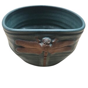 May include: A dark teal ceramic bowl with a rounded, slightly irregular rim. The exterior features a raised, embossed dragonfly design in a contrasting reddish-brown color, centered on the front of the bowl. The surface has a matte texture.