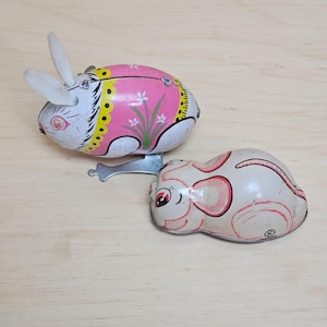 May include: Two vintage wind-up toys: a pink and white rabbit with white ears and a mouse with red accents. The rabbit has a yellow and black trim and floral details. The mouse has red ears and a tail. Both toys are made of metal.