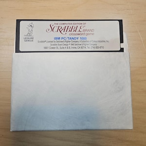 May include: A black 5.25-inch floppy disk for the computer edition of the Scrabble brand crossword game for IBM PC and Tandy 1000. The label features the Leisure Genius logo, game title, and company address in Irvine, CA, inside a white sleeve.