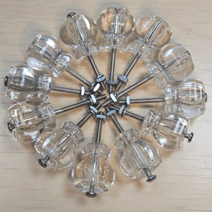 Puede incluir: Un conjunto de pomos de cristal transparente para armarios con un diseño decorativo acanalado. Cada pomo tiene un tornillo plateado y está dispuesto en un patrón circular sobre una superficie de madera clara. Los pomos miden unos 3,8 cm de diámetro.
