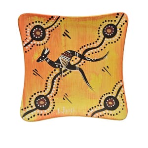 May include: Square, hand-painted ceramic dish with an Aboriginal art design. The dish features a kangaroo in black and white, surrounded by dotted lines and circular patterns on a yellow and orange background. The artist's signature is visible.