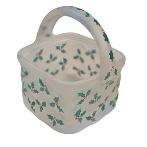 May include: A square-shaped white basket with a single arched handle, featuring a repeating pattern of green holly leaves and small red berries painted across the exterior and the handle, with a textured basket-weave surface design.