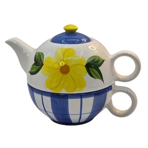 May include: A ceramic tea for one set featuring a white teapot with a yellow flower and green leaves, topped with a blue lid and a yellow knob. The base is a cup with a blue and white plaid pattern, featuring a circular handle on the side.