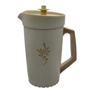 May include: A light beige plastic pitcher featuring a tan floral pattern on the front. The pitcher includes a matching lid with a central knob, a side handle, and a pouring spout. The base of the pitcher has a vertical ribbed texture design.
