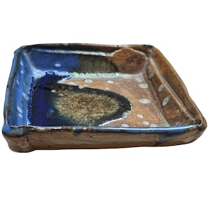 May include: A square ceramic dish with a hand-formed, uneven rim. The interior features a deep blue glaze on one side that transitions into a mottled brown and black center, accented by small white speckles across the tan, textured clay surface.