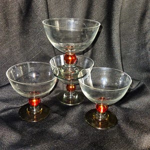 May include: A set of three clear glass coupes featuring a wide, shallow bowl and a stem accented with a single, spherical, metallic orange-red bead. Each glass rests on a circular base, showcasing a clean, transparent design with vibrant color.
