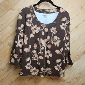 May include: A brown three-quarter sleeve top with a floral pattern in beige and cream. The top has a scoop neck and is hanging on a black hanger. The brand name is visible on the inner tag.