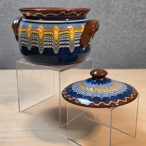 May include: A ceramic pot with two handles and a matching lid, featuring a dark blue base with a decorative marbled pattern in yellow, orange, and white. The rim and handles are solid brown, and the lid has a central knob with a matching design.