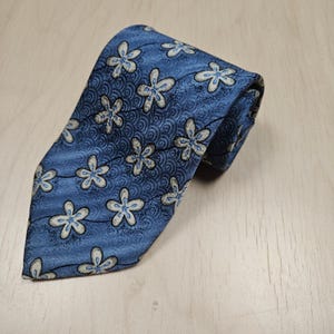 Puede incluir: Una corbata azul con un patrón floral repetido. Las flores son blancas con detalles grises, sobre un fondo azul texturizado. La corbata está doblada, mostrando el diseño y la tela.