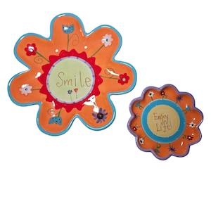 Puede incluir: Dos platos decorativos con forma de flor. El plato más grande es naranja con borde azul y la palabra "Smile" en el centro. El plato más pequeño es naranja con borde morado y las palabras "Enjoy Life!"