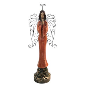 May include: A tall, standing angel figurine with a carved, reddish-brown wooden body, dark hair, and hands pressed together in prayer. The figure features black wire wings with a swirling pattern and a thin wire halo above the head, set on a dark base.
