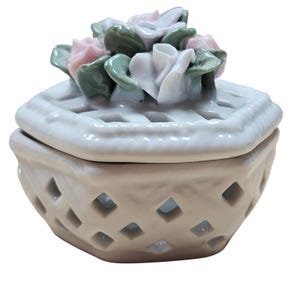May include: A white ceramic trinket box with a lattice design and a lid adorned with a cluster of pink, white, and green ceramic flowers. The box has a square shape with diamond-shaped cutouts.