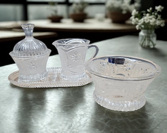 May include: A clear glass serving set with a sugar bowl, creamer, and bowl, all with floral etched designs. The set sits on a silver tray. The bowl has a label on the side. The set is on a gray surface.