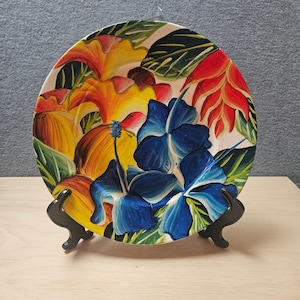 May include: A round ceramic plate featuring a vibrant hand-painted tropical floral design. The artwork displays large, bold blue hibiscus flowers, bright yellow and orange blossoms, and red tropical petals set against green foliage.