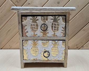 Könnte beinhalten: Kleiner Nachttisch aus Holz mit zwei Schubladen und einem Used-Look. Die Schubladen haben ein weißes und goldenes Ananas-Design und schwarze Ringgriffe. Der Nachttisch ist aus hellem Holz gefertigt und hat eine rustikale Ästhetik.