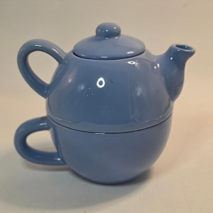 May include: A light blue ceramic tea set for one, featuring a stackable design with a teapot that sits securely on top of a matching teacup. The teapot has a rounded body, a curved handle, a short spout, and a lid with a small knob handle.