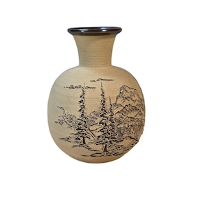 May include: A round, tan ceramic vase with a flared neck and a dark brown rim. The body of the vase features a dark, etched landscape design depicting tall pine trees and mountain peaks, creating a textured, detailed scene on the light surface.