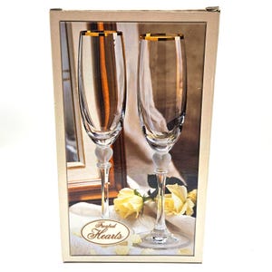 May include: A box set of two champagne flutes. The clear glass flutes have gold rims and heart-shaped bases. The box has a cream-colored background with the words "Frosted Hearts" in an oval design.