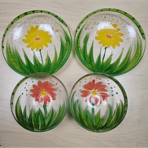 May include: Four clear glass bowls with a floral design. Two bowls feature a yellow flower, and two have a red flower. Each bowl has green grass and a green rim with colorful dots.