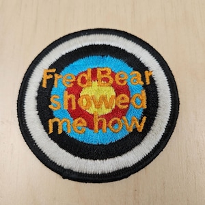 May include: A circular embroidered patch featuring a target design with concentric rings of yellow, red, blue, black, and white. The text "Fred Bear showed me how" is stitched in gold thread across the centre of the target design.