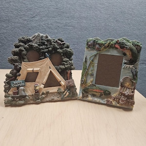 May include: Two decorative resin picture frames. The left frame features a camping scene with a tent, trees, a bear, and a sign reading HAPPY CAMPERS. The right frame depicts a fishing scene with a rod, a trout, and a woven creel basket.