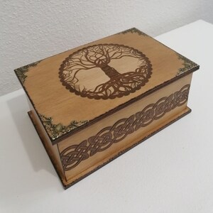 Puede incluir: Una caja rectangular de madera con acabado marrón claro. La tapa presenta un diseño detallado de árbol de la vida, y los lados están adornados con un patrón de nudo celta. Los acentos de esquina de latón antiguo añaden un toque decorativo.