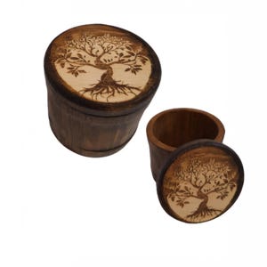 Op de afbeelding: Twee houten dozen, waarvan één open, met deksels met een lichtgekleurd boomontwerp dat in het hout is geëtst. De cilindrische dozen zijn gemaakt van donkerbruin hout. De open doos toont het interieur.