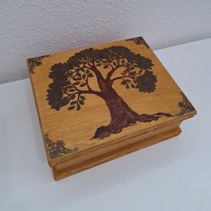 Op de afbeelding: Houten kist met een gedetailleerd boomontwerp op het deksel. De boom is bruin met groene bladeren, tegen een lichtbruine achtergrond. Decoratieve metalen hoekaccenten. Een handgemaakte opbergdoos.