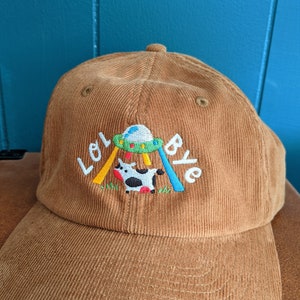 May include: A brown corduroy baseball cap with an embroidered design of a cow being abducted by a UFO. The text "Lol Bye" is embroidered on the cap.