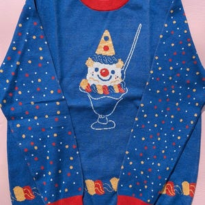 May include: A blue long-sleeve crewneck jumper with a red collar, cuffs, and hem. The front features a graphic of a clown in a party hat inside a sundae glass. The sleeves have a pattern of small red and yellow dots, and the label reads X-LARGE.