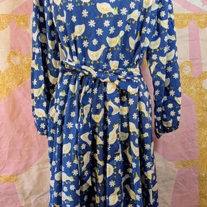 May include: A blue dress with a white duck and flower pattern. The long-sleeved dress has a tie waist and a boat neckline. The dress is displayed on a mannequin.