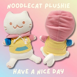 May include: White plush cat wearing a pink hoodie with a yellow and blue shirt that says "Have a nice day". The cat is sitting on a pink and blue background.