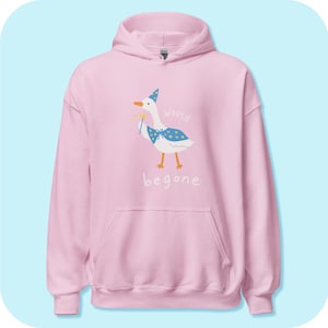 May include: Pink hooded sweatshirt featuring a cartoon illustration of a goose wearing a wizard hat and holding a wand. The goose is saying "woosh" and "begone".