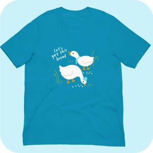 May include: A turquoise blue t-shirt featuring a white graphic of two geese. The geese are looking down at a pile of white dots. The text "Let's get this bread" is written in white above the geese.