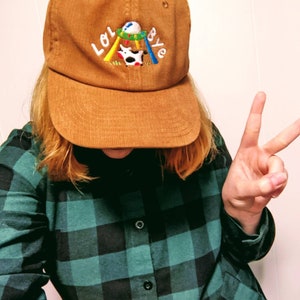 May include: A brown corduroy baseball cap with an embroidered design featuring a cow on a swing set and the text "LOL BYE".