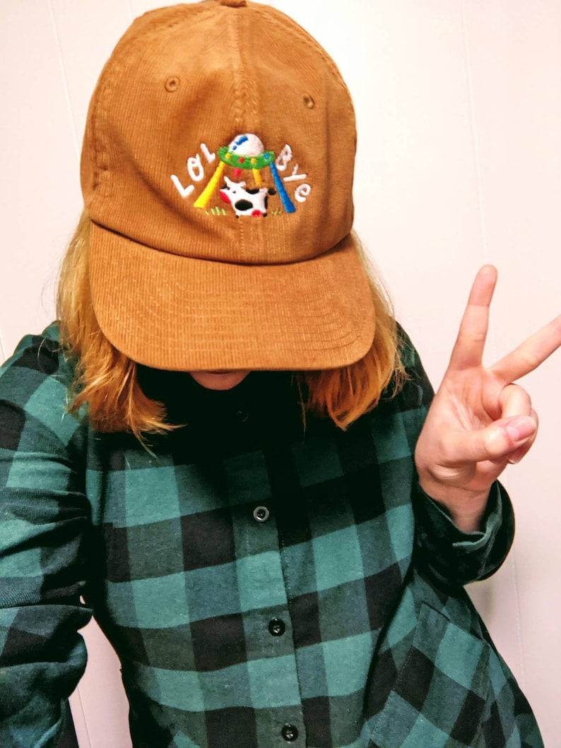 May include: A brown corduroy baseball cap with an embroidered design featuring a cow on a swing set and the text "LOL BYE".