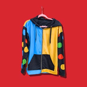 May include: A zip-up hoodie featuring a colour-blocked design with a bright blue right side and a yellow left side. The sleeves are black with large, multicoloured polka dots. It includes a black hood, black drawstrings, and a black front pocket.