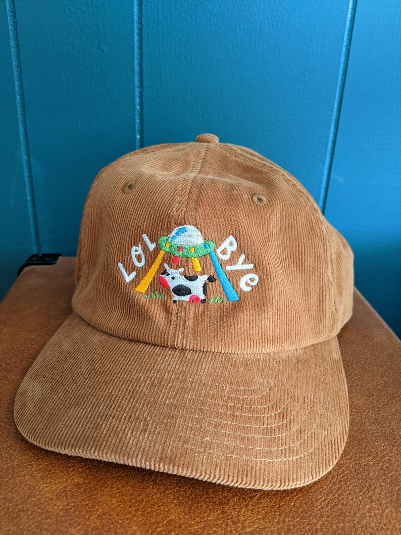 May include: A brown corduroy baseball cap with an embroidered design of a cow being abducted by a UFO. The text "Lol Bye" is embroidered on the cap.