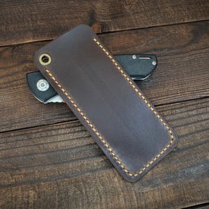 May include: A dark brown leather knife sheath with a brass grommet and tan stitching. A black folding knife is partially visible. The sheath is rectangular and appears to be handmade. The background is a dark wooden surface.