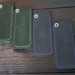 May include: Four leather keychains in shades of green and blue, arranged on a dark wooden surface. Two green keychains have white stitching, and two blue keychains have blue stitching. The word "SPOOKY" is embossed on two of the keychains.