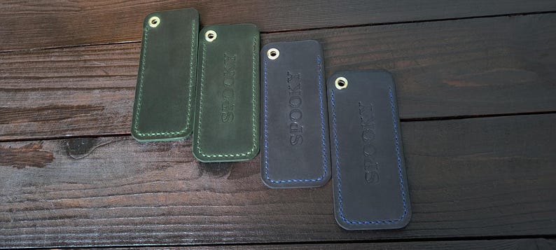 May include: Four leather keychains in shades of green and blue, arranged on a dark wooden surface. Two green keychains have white stitching, and two blue keychains have blue stitching. The word "SPOOKY" is embossed on two of the keychains.