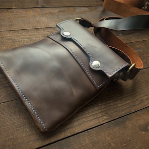 Könnte beinhalten: Eine braune Ledertasche mit Klappenverschluss und blauen Nähten. Die Tasche hat einen langen verstellbaren Riemen und eine Metallschnalle.