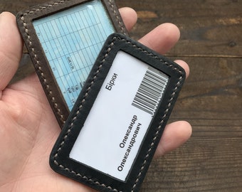 May include: Two leather ID card holders with stitched edges. The top holder is brown with a clear plastic window and a lined grid. The bottom holder is black with a clear plastic window and the text "Бирки Олександр Олександрович" in white.