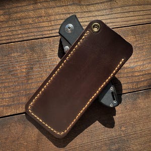May include: A brown leather knife sheath with a contrasting stitched border and a brass grommet. The sheath is shown on a wooden surface, partially concealing a black folding knife.